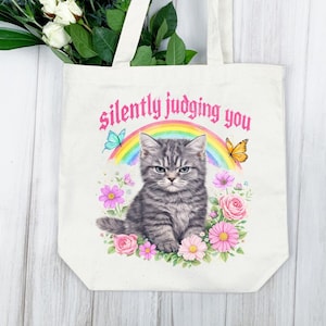 Pu&ograve; includere: Una borsa tote color crema con un'illustrazione ad acquerello di un gatto tabby grigio con un'espressione non impressionata. Il gatto &egrave; circondato da fiori, farfalle e un arcobaleno. Il testo "silently judging you" &egrave; arcuato sopra il gatto.