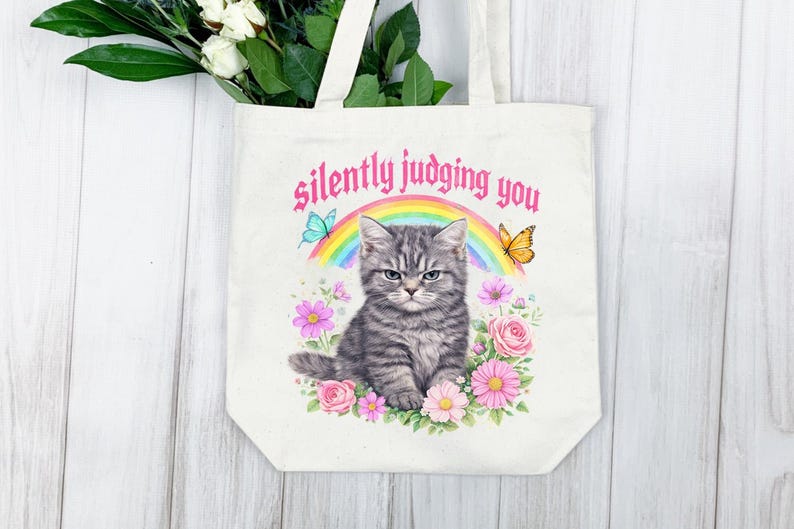Pu&ograve; includere: Una borsa tote color crema con un'illustrazione ad acquerello di un gatto tabby grigio con un'espressione non impressionata. Il gatto &egrave; circondato da fiori, farfalle e un arcobaleno. Il testo "silently judging you" &egrave; arcuato sopra il gatto.