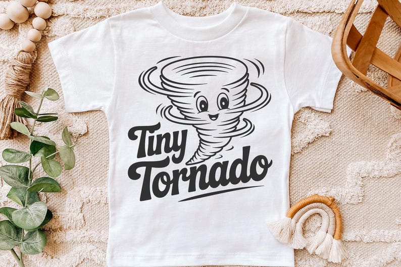 Peut inclure: T-shirt blanc avec un dessin anim&eacute; en noir et blanc d'une tornade souriante. Le texte "Tiny Tornado" est &eacute;crit en dessous dans une police de caract&egrave;res audacieuse et amusante. Un design amusant pour les enfants.
