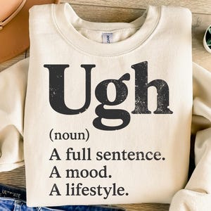 May include: Cream-colored sweatshirt with the word "Ugh" in distressed black lettering. Below, the definition reads: "(noun) A full sentence. A mood. A lifestyle."