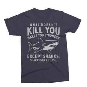 May include: Navy blue t-shirt with the text "WHAT DOESN'T KILL YOU MAKES YOU STRONGER EXCEPT SHARKS. SHARKS WILL KILL YOU." A white shark illustration is centered below the text. The shirt is a crew neck.