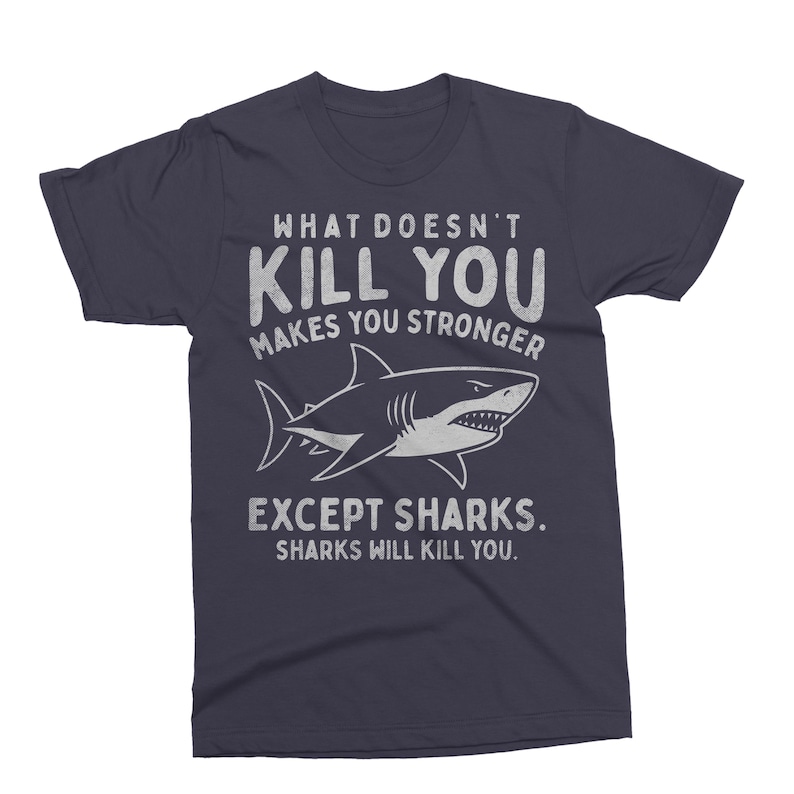 May include: Navy blue t-shirt with the text "WHAT DOESN'T KILL YOU MAKES YOU STRONGER EXCEPT SHARKS. SHARKS WILL KILL YOU." A white shark illustration is centered below the text. The shirt is a crew neck.