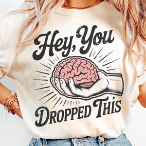 May include: Cream-colored t-shirt with the text "Hey, You Dropped This" in black. The graphic features a hand holding a pink brain, with radiating lines. The design has a vintage, distressed look.