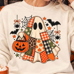 Puede incluir: Una sudadera color crema con un gráfico central de un fantasma hecho de varios patrones de patchwork, incluyendo cuadros, estampado de leopardo y lunares. El fantasma sostiene un cubo de calabaza naranja, rodeado de arañas, telarañas y murciélagos.