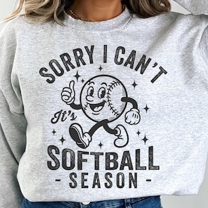 Op de afbeelding: Een lichtgrijs sweatshirt met de tekst "SORRY I CAN'T It's SOFTBALL SEASON" in het zwart. De afbeelding toont een cartoon honkbalpersonage dat een duim omhoog geeft. Het sweatshirt heeft een casual, sportieve stijl.