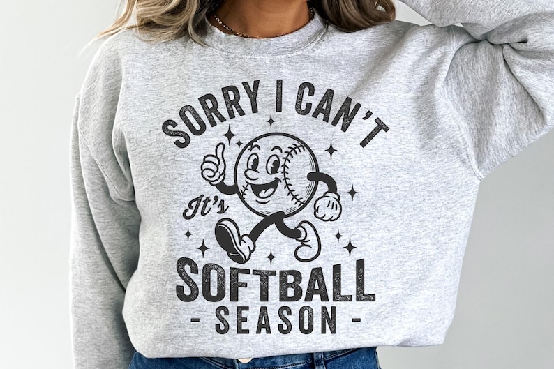Op de afbeelding: Een lichtgrijs sweatshirt met de tekst "SORRY I CAN'T It's SOFTBALL SEASON" in het zwart. De afbeelding toont een cartoon honkbalpersonage dat een duim omhoog geeft. Het sweatshirt heeft een casual, sportieve stijl.