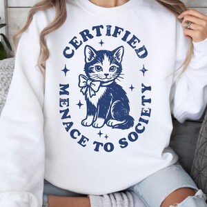 May include: White sweatshirt with a navy blue graphic of a cat wearing a bow tie, surrounded by the text "CERTIFIED MENACE TO SOCIETY". The design includes stars and a distressed, vintage aesthetic.