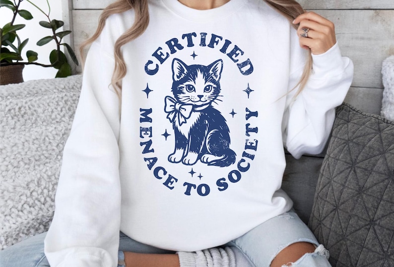May include: White sweatshirt with a navy blue graphic of a cat wearing a bow tie, surrounded by the text "CERTIFIED MENACE TO SOCIETY". The design includes stars and a distressed, vintage aesthetic.