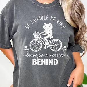 May include: Dark grey t-shirt featuring a white graphic of a frog on a bicycle with a flower basket. The text says "BE HUMBLE BE KIND" and "leave your worries BEHIND".