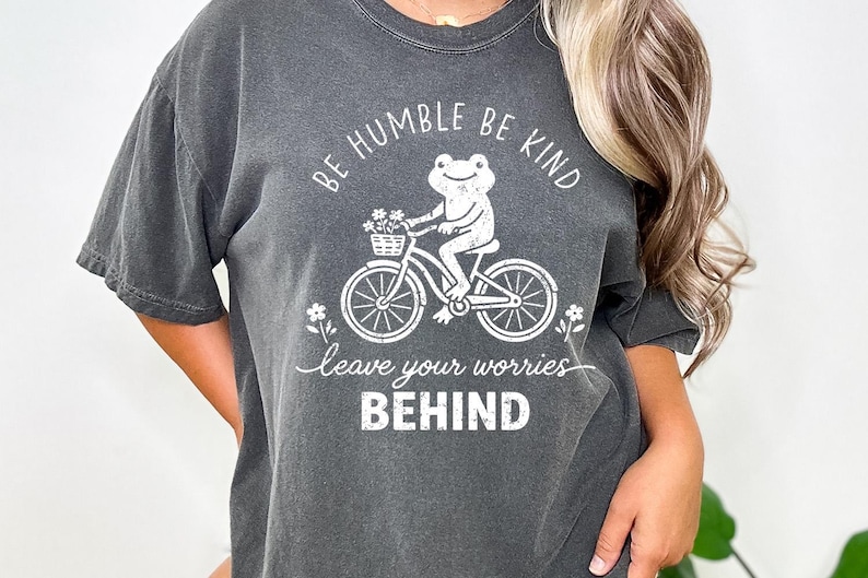 May include: Dark grey t-shirt featuring a white graphic of a frog on a bicycle with a flower basket. The text says "BE HUMBLE BE KIND" and "leave your worries BEHIND".