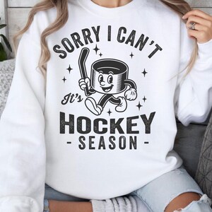 May include: White sweatshirt with the text "SORRY I CAN'T It's HOCKEY SEASON" in black. The graphic features a cartoon hockey puck with a hockey stick. The sweatshirt is a casual, comfortable style.
