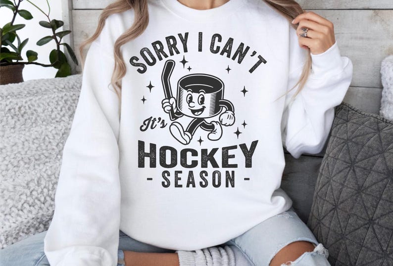 May include: White sweatshirt with the text "SORRY I CAN'T It's HOCKEY SEASON" in black. The graphic features a cartoon hockey puck with a hockey stick. The sweatshirt is a casual, comfortable style.