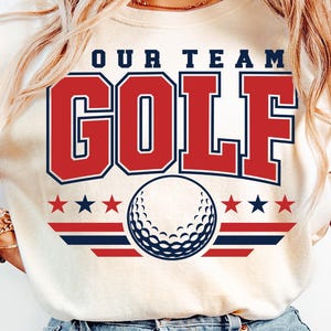 May include: Cream-colored t-shirt with the text "OUR TEAM GOLF" in red and blue. A golf ball graphic is centered below the text, with red and blue stars and stripes. The shirt is a casual style.