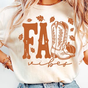 May include: Cream-colored t-shirt with a brown graphic that reads "Fall Vibes." The design includes a pair of cowboy boots, a pumpkin, and autumn leaves. The word "Fall" is in a bold, western-style font.