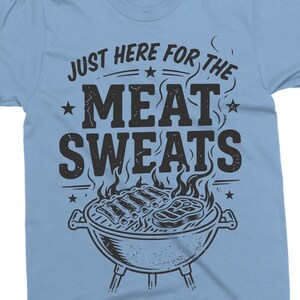May include: Light blue t-shirt with the phrase "JUST HERE FOR THE MEAT SWEATS" in distressed black lettering. A graphic of a grill with ribs and steak is below the text. The shirt is short-sleeved.
