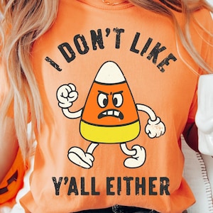 May include: Orange t-shirt with the text "I DON'T LIKE Y'ALL EITHER" and a cartoon candy corn character with an angry expression. The candy corn is orange, yellow, and white.