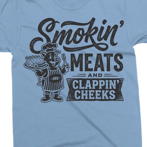 May include: Light blue t-shirt with the text "Smokin' MEATS and CLAPPIN' CHEEKS". The shirt features a cartoon pig chef holding a platter of ribs and a fork. The design is in black.