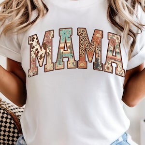 Op de afbeelding: Wit T-shirt met het woord "MAMA" in een decoratief, gebogen ontwerp. De letters zijn gevuld met verschillende patronen, waaronder koeienhuid, woestijnlandschappen en luipaardprint. Het is een casual kledingstuk voor dagelijks gebruik.