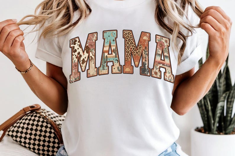 Op de afbeelding: Wit T-shirt met het woord "MAMA" in een decoratief, gebogen ontwerp. De letters zijn gevuld met verschillende patronen, waaronder koeienhuid, woestijnlandschappen en luipaardprint. Het is een casual kledingstuk voor dagelijks gebruik.
