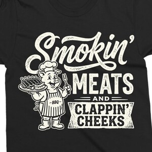 May include: Black t-shirt with a white graphic. The graphic features a cartoon pig chef holding a plate of ribs and a fork, with the words "Smokin' MEATS and CLAPPIN' CHEEKS" in a vintage style.