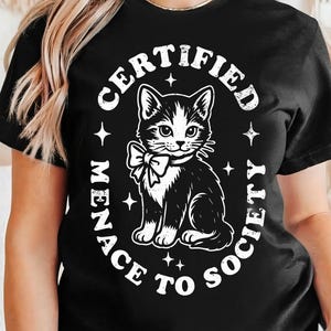 May include: Black t-shirt featuring a black and white cat illustration with a bow tie. The text reads "CERTIFIED MENACE TO SOCIETY" in a circular design around the cat. The design includes star accents.