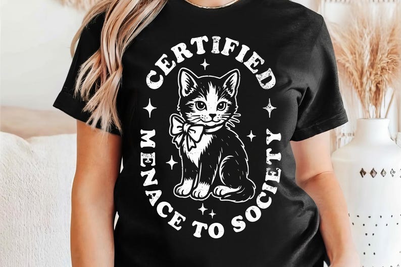 May include: Black t-shirt featuring a black and white cat illustration with a bow tie. The text reads "CERTIFIED MENACE TO SOCIETY" in a circular design around the cat. The design includes star accents.