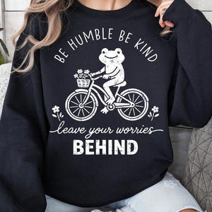 May include: Black sweatshirt featuring a white graphic of a frog on a bicycle with a flower basket. The text says "Be Humble Be Kind" and "leave your worries BEHIND".
