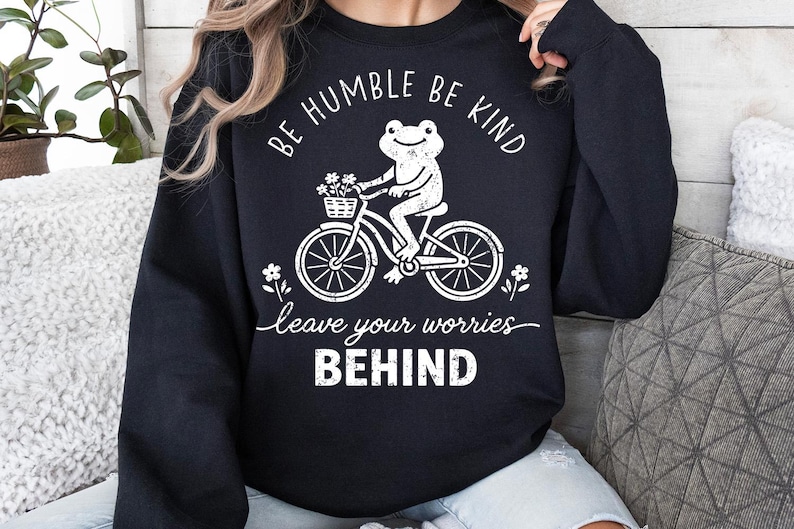 May include: Black sweatshirt featuring a white graphic of a frog on a bicycle with a flower basket. The text says "Be Humble Be Kind" and "leave your worries BEHIND".
