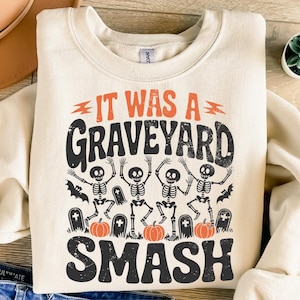 May include: Cream-colored sweatshirt with the text "IT WAS A GRAVEYARD SMASH" in black and orange. The graphic features dancing skeletons, bats, tombstones, and pumpkins, perfect for Halloween.
