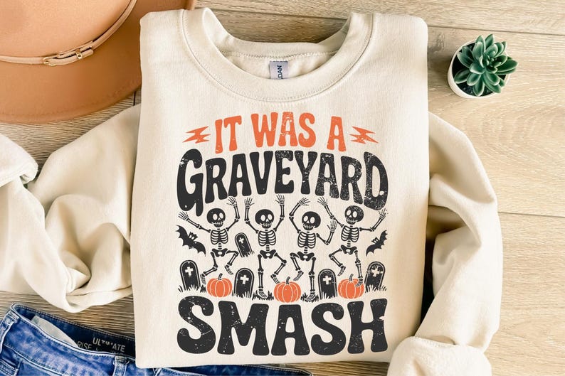 May include: Cream-colored sweatshirt with the text "IT WAS A GRAVEYARD SMASH" in black and orange. The graphic features dancing skeletons, bats, tombstones, and pumpkins, perfect for Halloween.