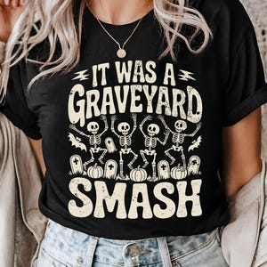May include: Black t-shirt with the text "IT WAS A GRAVEYARD SMASH" in a distressed font. The graphic features dancing skeletons, pumpkins, and tombstones, perfect for Halloween.