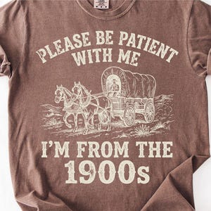 May include: Brown t-shirt with the text "PLEASE BE PATIENT WITH ME" above an illustration of a covered wagon pulled by horses. Below the illustration is the text "I'M FROM THE 1900s".