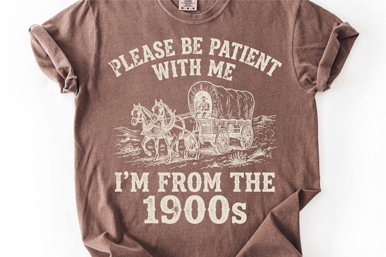 May include: Brown t-shirt with the text "PLEASE BE PATIENT WITH ME" above an illustration of a covered wagon pulled by horses. Below the illustration is the text "I'M FROM THE 1900s".