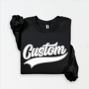 May include: Black crewneck sweatshirt with the word "Custom" in a white, cursive font with a white banner underneath. The sweatshirt is folded, with the sleeves visible.