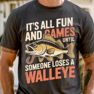 May include: Dark gray t-shirt with the text "IT'S ALL FUN AND GAMES UNTIL SOMEONE LOSES A WALLEYE." The shirt features a graphic of a walleye fish with a fishing hook. The text is in white and orange.