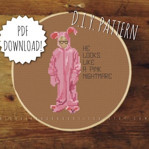 May include: A brown embroidery hoop with a cross-stitch pattern of a person in a pink bunny costume. The text "HE LOOKS LIKE A PINK NIGHTMARE" is stitched below the figure. The hoop has a white label that says "PDF DOWNLOAD!"