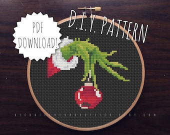 Può includere: Uno schema di punto croce della mano del Grinch che tiene un ornamento natalizio rosso. Lo schema è incorniciato in un cerchio di legno e il testo "DIY PATTERN" è scritto sopra il cerchio. Il testo "PDF DOWNLOAD!" è scritto in una forma a stella bianca a sinistra del cerchio.