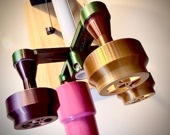 Peut inclure: Un accessoire de tournage de tasses imprimé en 3D multicolore avec un cadre central vert et quatre bras extensibles. Chaque bras supporte un adaptateur cylindrique en or métallique, violet foncé et rose uni, avec une tige centrale blanche.