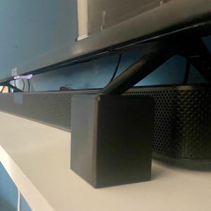 May include: A black soundbar with a speaker grille and two black rectangular supports. The soundbar is mounted on a white shelf below a black television.