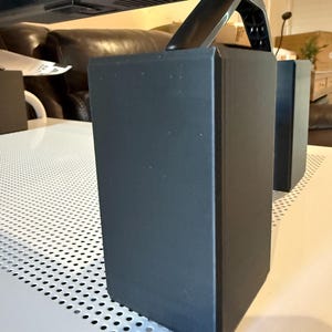 May include: Black monitor stand with three rectangular supports. The supports are positioned under the monitor, providing stability. The stand is on a white surface with a perforated design.