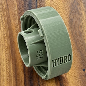 Könnte beinhalten: Ein zylindrischer Kunststoffadapter in Salbeigrün mit einem zentralen Rohr, das den geprägten Text 15S trägt, und einem Außenring mit dem geprägten Text HYDRO. Das Bauteil verfügt über ein Innengewinde und stützende Rippen zwischen den Teilen.