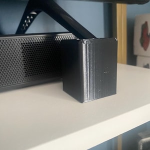 Puede incluir: Un soporte negro impreso en 3D para un televisor u otros dispositivos electrónicos. El soporte está en un estante blanco.