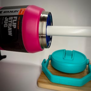 Peut inclure: Une carafe Stanley Flip Straw Jug rose avec une paille blanche et un couvercle turquoise. La carafe porte l'inscription "THE ICEFLOW" et "FLIP STRAW JUG" sur le côté.