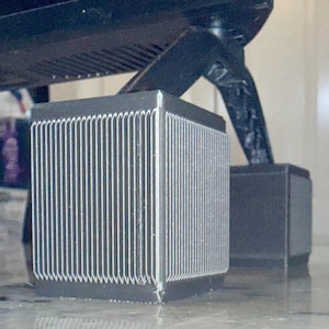 May include: Close-up of two black, cube-shaped objects with vertical, silver lines on the sides. The objects appear to be supporting a larger, black structure above them. The objects are on a reflective surface.