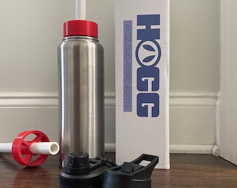 Peut inclure: Une bouteille hydro en acier inoxydable de 1183 ml avec un bouchon rouge et une paille blanche, accompagnée de sa boîte blanche étiquetée HOGG 40 OZ. DUO HYDRO BOTTLE. Un embout de paille rouge et un couvercle noir avec poignée sont inclus.