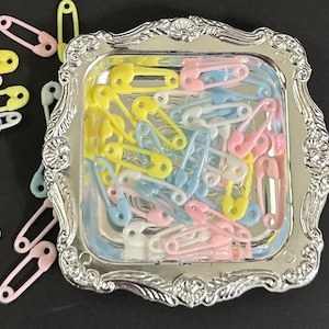 May include: A silver tray filled with pastel blue, pink, and yellow safety pins. The tray has a decorative, ornate border.
