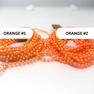 May include: Two coiled strands of bright orange ball chain, labeled ORANGE #1 and ORANGE #2. Each strand consists of small, uniform, spherical beads connected by thin metal links, arranged in loose, overlapping loops on a plain white surface.