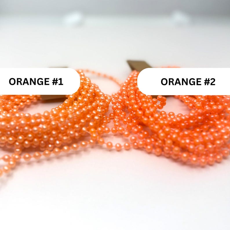 May include: Two coiled strands of bright orange ball chain, labeled ORANGE #1 and ORANGE #2. Each strand consists of small, uniform, spherical beads connected by thin metal links, arranged in loose, overlapping loops on a plain white surface.