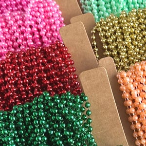 May include: Multiple strands of ball chain necklaces in various colors including hot pink, deep red, emerald green, metallic gold, light mint green, and peach, each neatly coiled and displayed on individual rectangular brown cardboard cards.