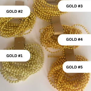 May include: Five coils of metallic gold ball chains, each secured with a tan paper tag. The coils are labeled with black text as GOLD #1, GOLD #2, GOLD #3, GOLD #4, and GOLD #5. The chains consist of small, uniform spherical beads linked together.